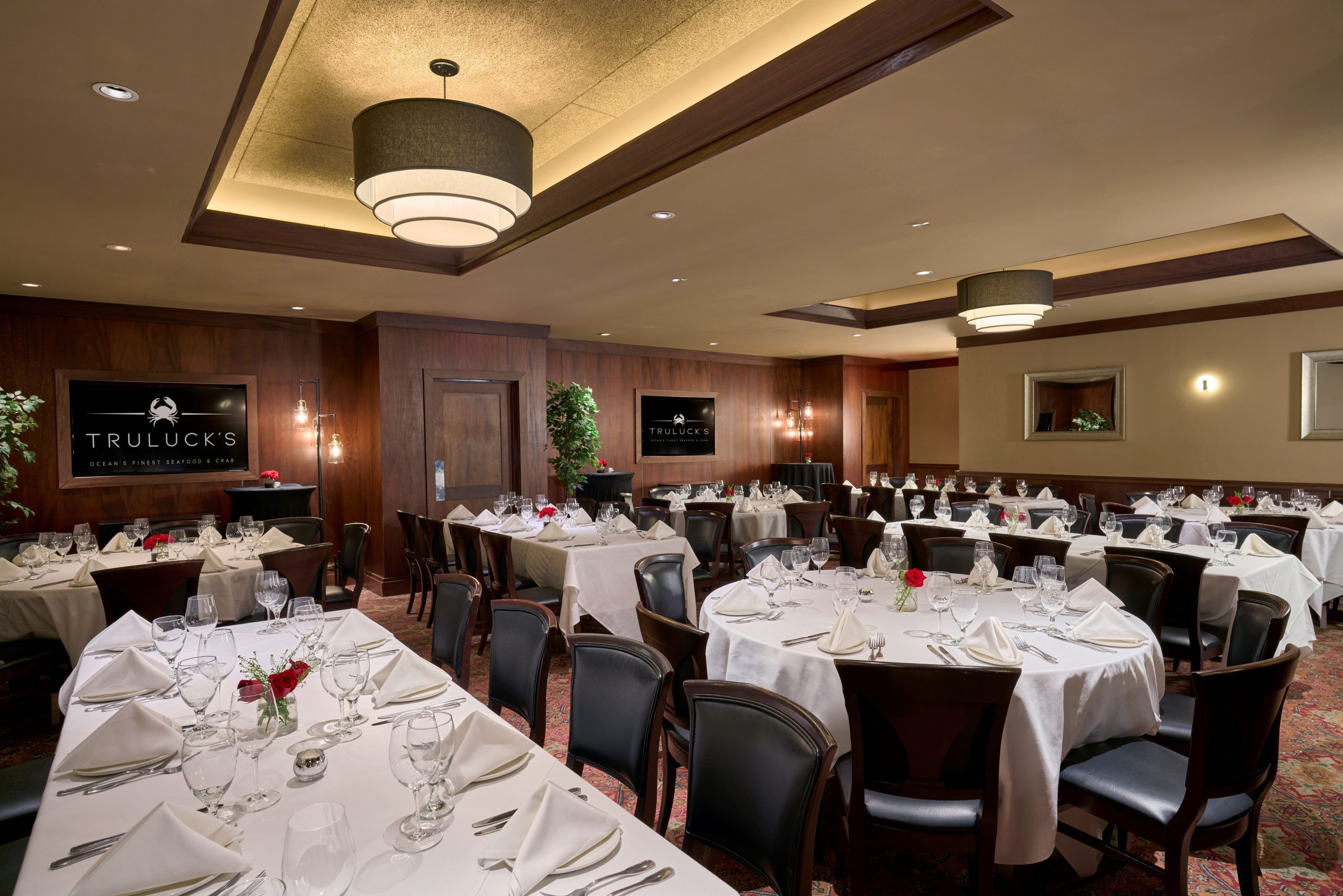 Naples rooms 1 and 2, a spacious private dining room at Truluck’s with multiple round and rectangular tables draped in white linens, set with polished glassware, silverware, and folded napkins. The room features warm wood-paneled walls, soft lighting, modern chandeliers, wall-mounted Truluck’s branding, and a refined, upscale ambiance suitable for large gatherings.