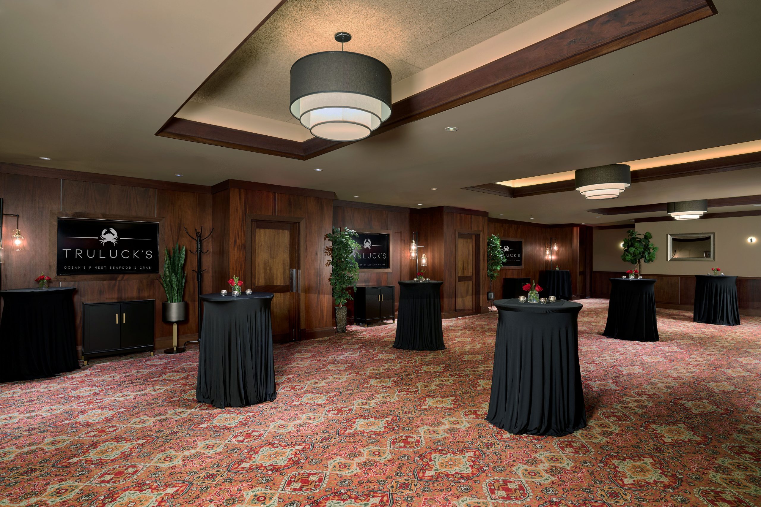 Naples 1, 2 and 3 - an elegant event space at Truluck's, featuring several high-top tables with black tablecloths, small floral centerpieces with red roses, and candles. The room has dark wood-paneled walls, a patterned red carpet, and modern lighting with large, tiered chandeliers. Decor includes potted plants, a mirror, and wall sconces, creating a sophisticated ambiance. Signs on the wall read "Truluck's Ocean's Finest Seafood & Crab."