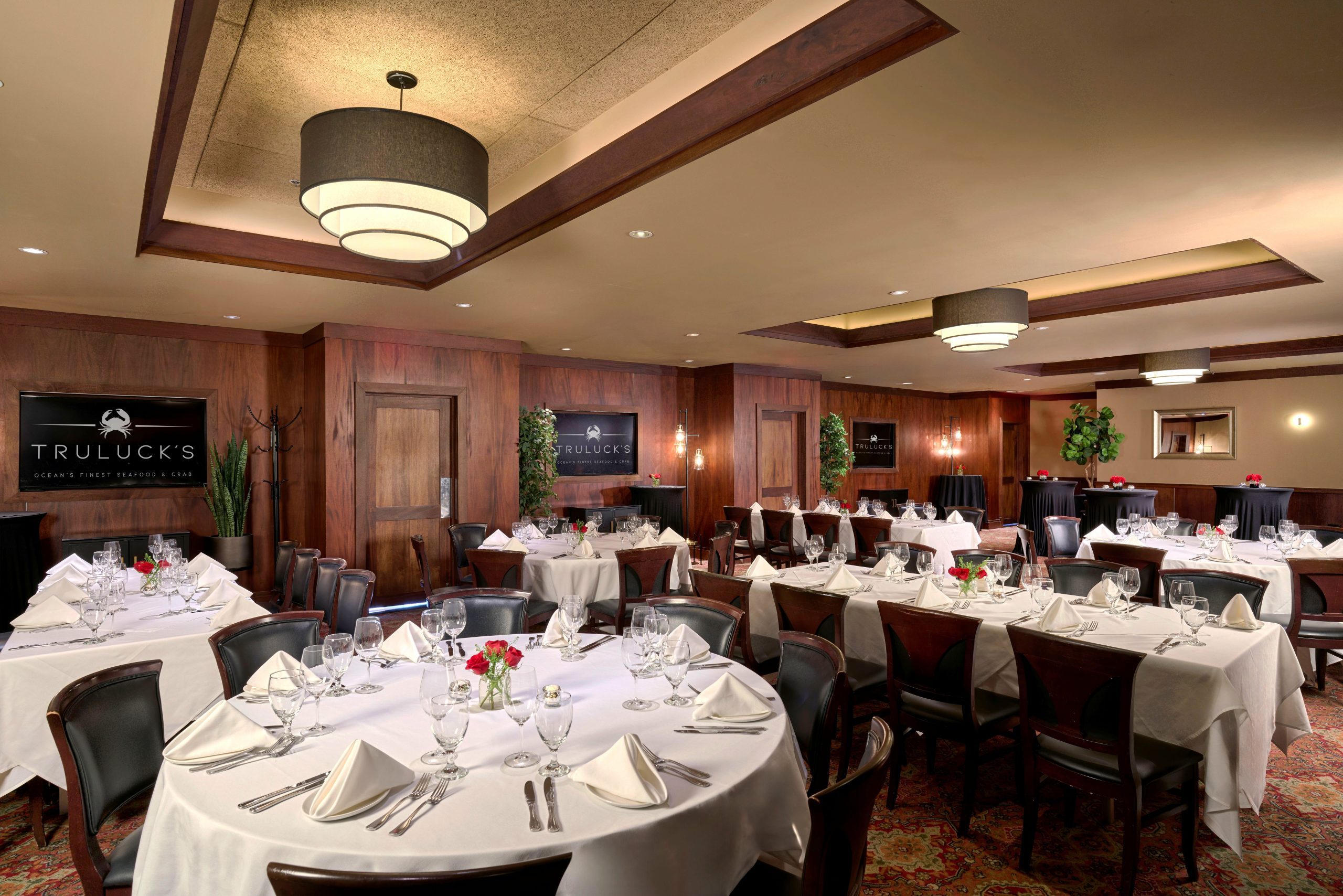 Naples room 1, 2 and 3 - a sophisticated dining room at Truluck's, featuring a mix of round tables and long rectangular tables, set with white tablecloths, glassware, and small floral centerpieces with red roses. The room has dark wood-paneled walls, a patterned red carpet, and modern lighting with a large, tiered chandelier. Decor includes potted plants, mirrors, and wall sconces, creating an elegant ambiance. A sign on the wall reads "Truluck's Ocean's Finest Seafood & Crab."