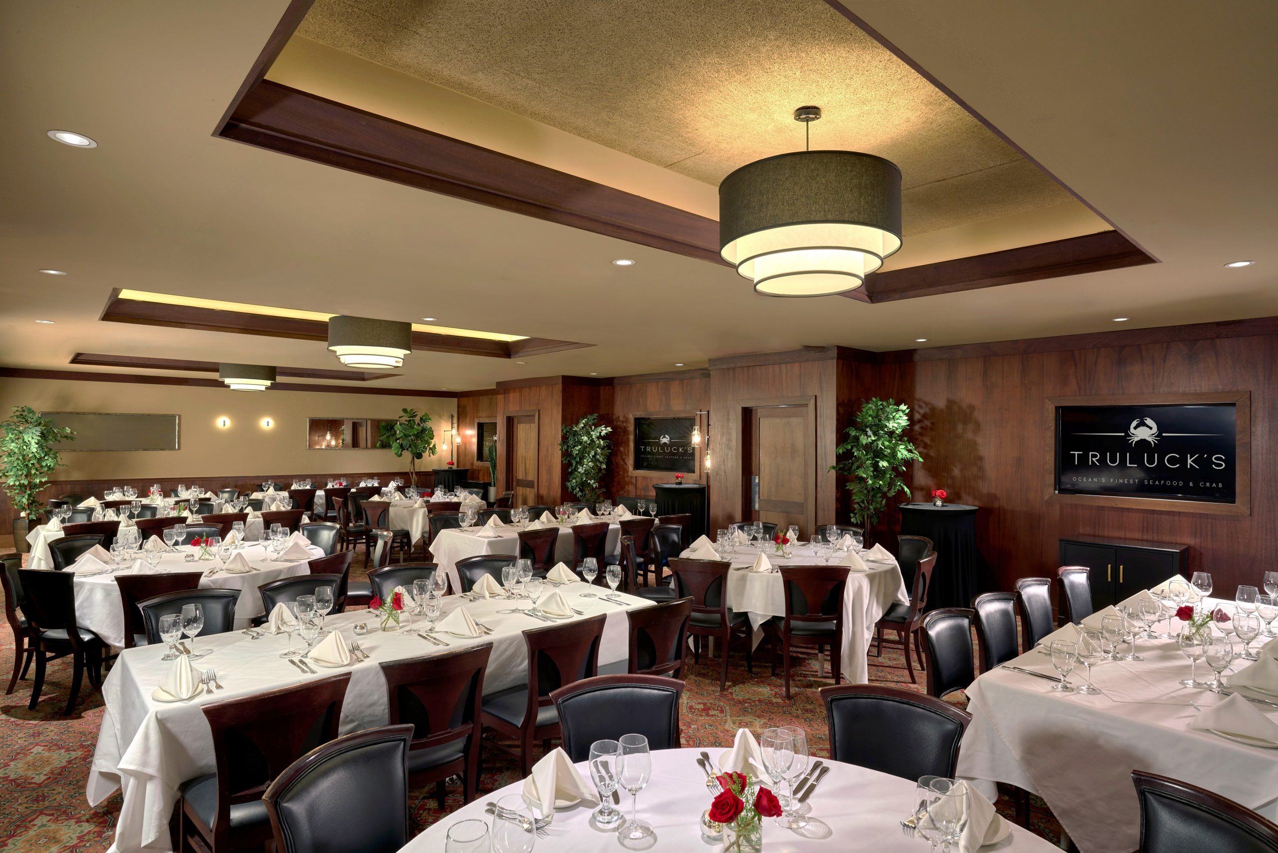 Naples room 1, 2 and 3 - a large elegant dining room at Truluck's, featuring a mix of round tables and long rectangular tables, set with white tablecloths, glassware, and small floral centerpieces with red roses. The room has dark wood-paneled walls, a patterned red carpet, and modern lighting with a large, tiered chandelier. Decor includes potted plants, mirrors, and wall sconces, creating an elegant ambiance. A sign on the wall reads "Truluck's Ocean's Finest Seafood & Crab."