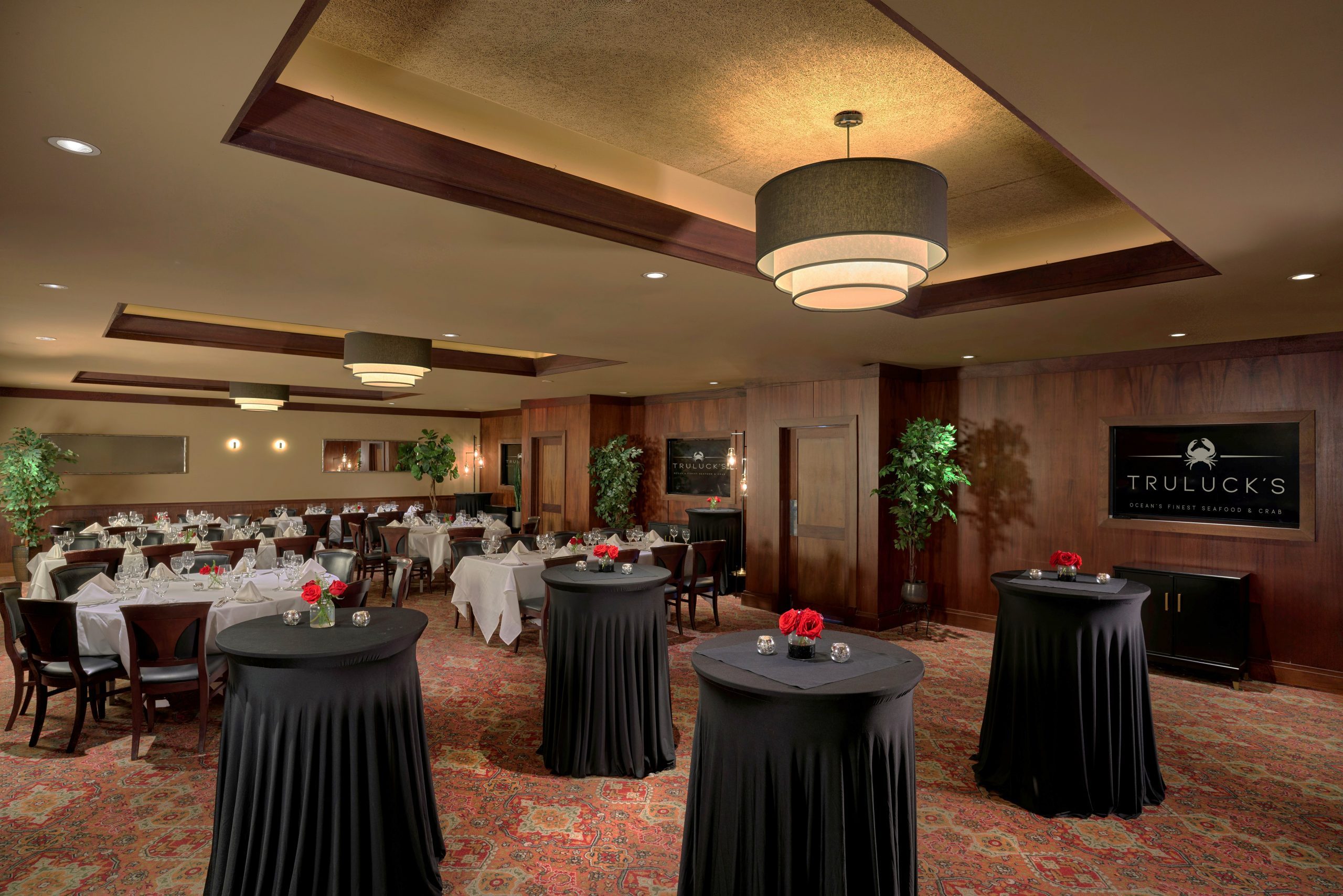 Naples room 1, 2 and 3 - a sophisticated dining room at Truluck's, featuring a mix of round tables and long rectangular tables, set with white tablecloths, glassware, and small floral centerpieces with red roses. Also pictured are 4 tall high boy tables draped in wavy black linens with small floral centerpieces and candles. The room has dark wood-paneled walls, a patterned red carpet, and modern lighting with a large, tiered chandelier. Decor includes potted plants, mirrors, and wall sconces, creating an elegant ambiance. A sign on the wall reads "Truluck's Ocean's Finest Seafood & Crab."