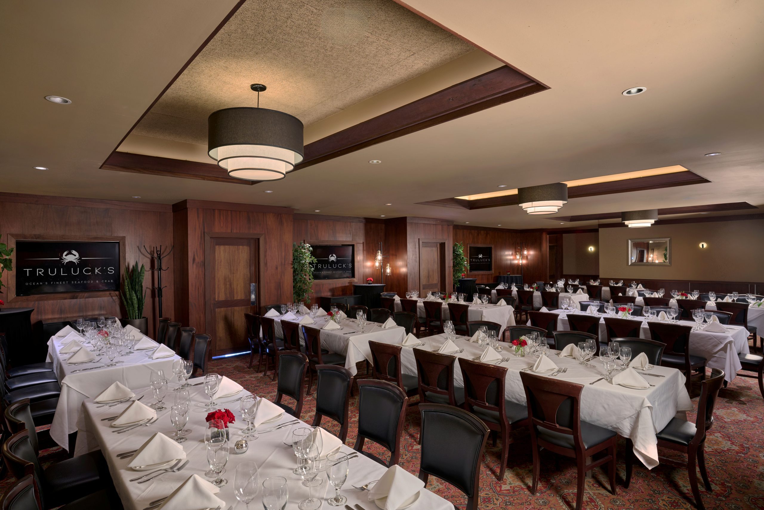 Naples room 1, 2 and 3 - a large refined private dining room at Truluck's, featuring long rectangular tables, set with white tablecloths, glassware, and small floral centerpieces with red roses. The room has dark wood-paneled walls, a patterned red carpet, and modern lighting with a large, tiered chandelier. Decor includes potted plants, mirrors, and wall sconces, creating an elegant ambiance. A sign on the wall reads 