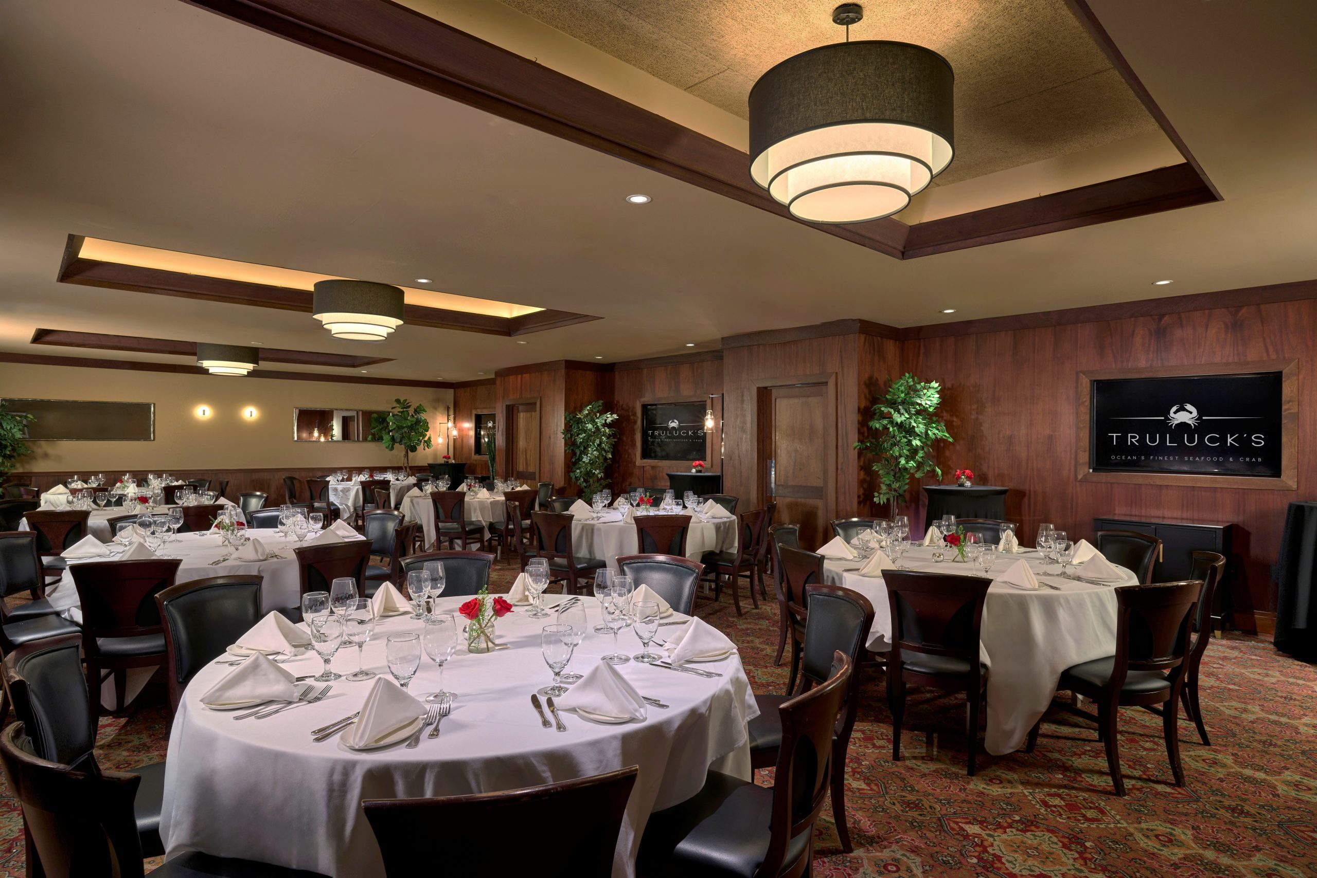 Naples room 1, 2 and 3 - a polished private dining room at Truluck's, featuring round tables set with white tablecloths, glassware, and small floral centerpieces with red roses. The room has dark wood-paneled walls, a patterned red carpet, and modern lighting with a large, tiered chandelier. Decor includes potted plants, mirrors, and wall sconces, creating an elegant ambiance. A sign on the wall reads "Truluck's Ocean's Finest Seafood & Crab."
