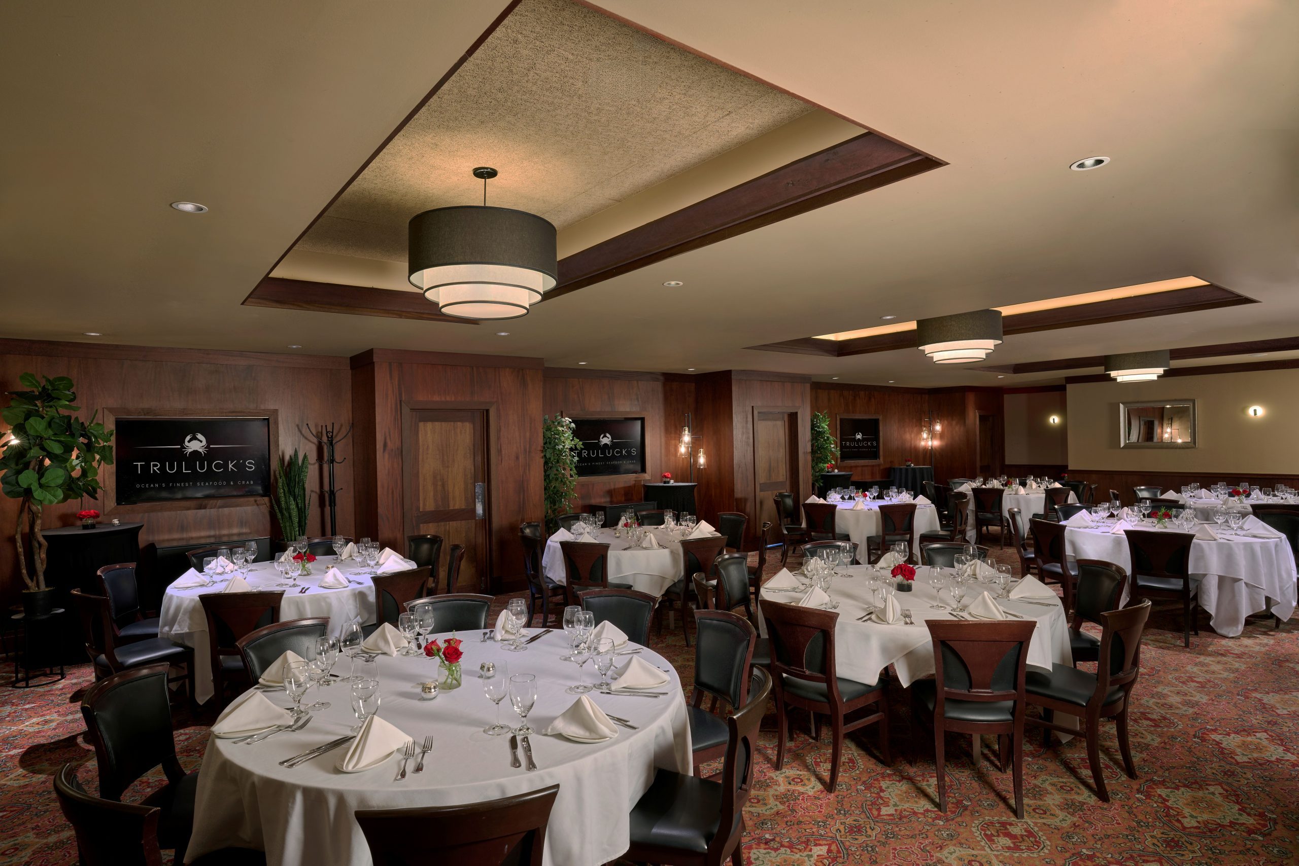 Naples room 1, 2 and 3 - a classy private dining room at Truluck's, featuring round tables set with white tablecloths, glassware, and small floral centerpieces with red roses. The room has dark wood-paneled walls, a patterned red carpet, and modern lighting with a large, tiered chandelier. Decor includes potted plants, mirrors, and wall sconces, creating an elegant ambiance. A sign on the wall reads "Truluck's Ocean's Finest Seafood & Crab."