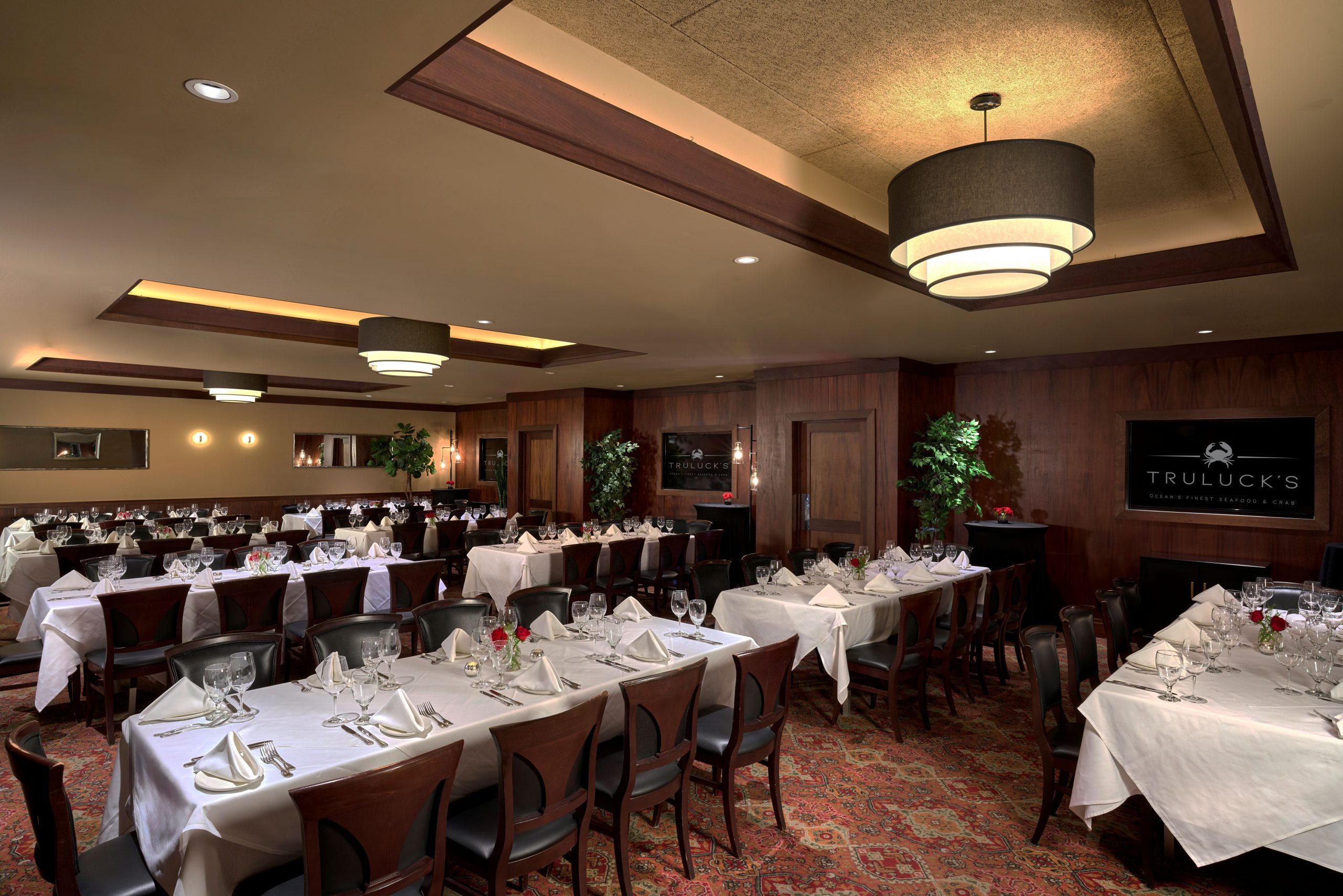 Naples room 1, 2 and 3 - a classy dining room at Truluck's, featuring long rectangular tables, set with white tablecloths, glassware, and small floral centerpieces with red roses. The room has dark wood-paneled walls, a patterned red carpet, and modern lighting with a large, tiered chandelier. Decor includes potted plants, mirrors, and wall sconces, creating an elegant ambiance. A sign on the wall reads "Truluck's Ocean's Finest Seafood & Crab."