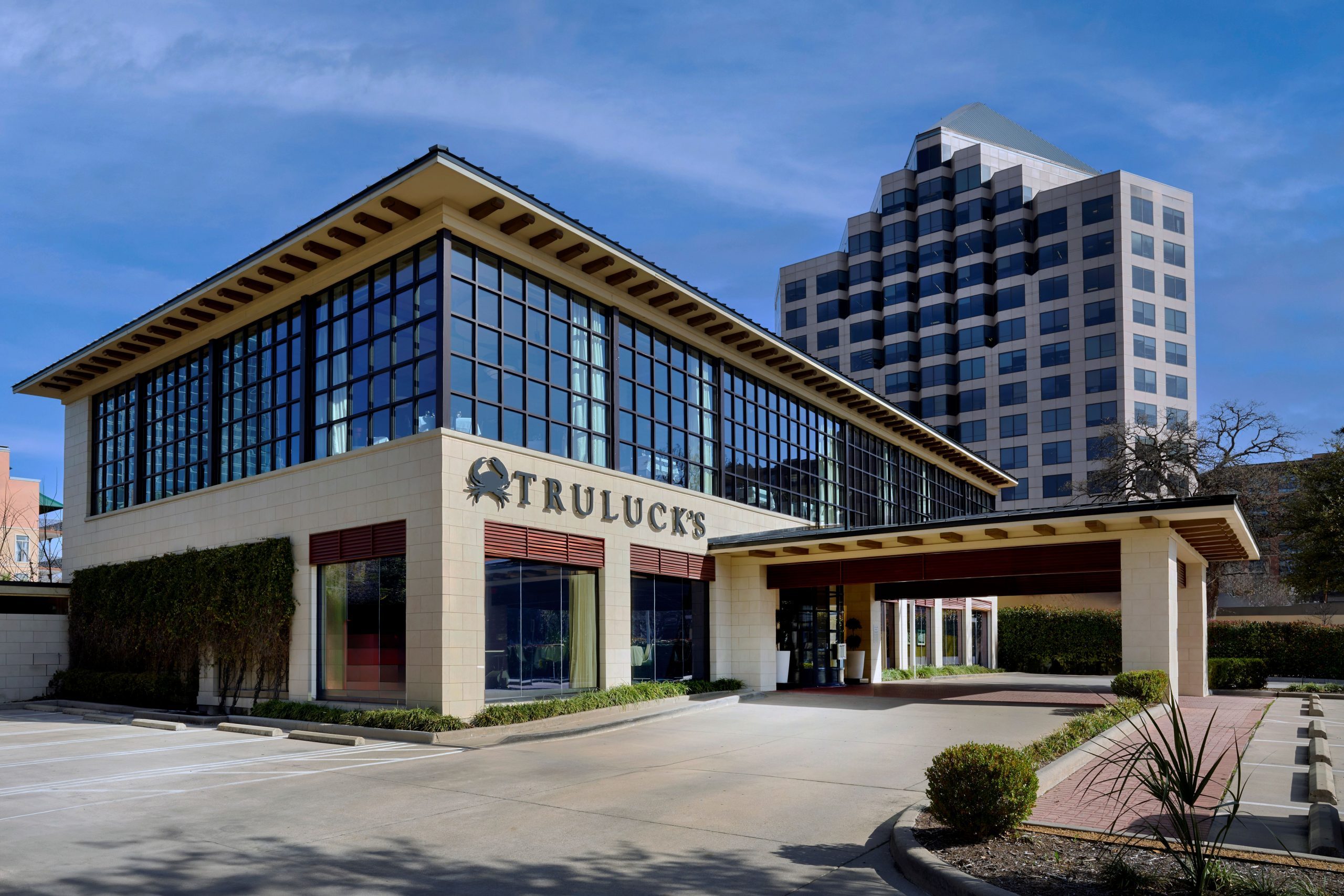 The exterior of Truluck's restaurant, featuring a modern building with large, grid-patterned windows and light-colored stone walls. The name "TRULUCK'S" is prominently displayed with a crab logo above the entrance. The entrance has a covered walkway, surrounded by neatly trimmed bushes and greenery. A tall, contemporary office building is visible in the background under a clear blue sky.