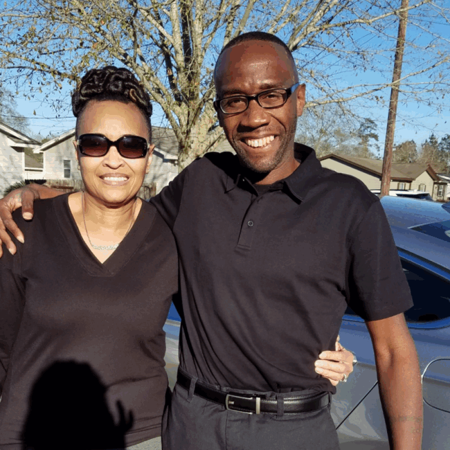 Dre Reed in a black polo style shirt and black slacks standing with his arm around his Mom's shoulder outside with a tree in the background.