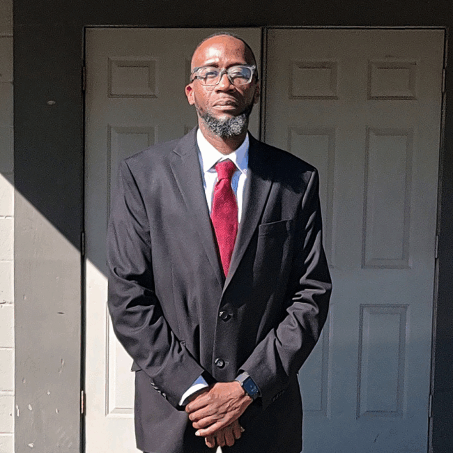 Dre Reed dressed in a black suite with a garnet colored tie and white dress shirt standing with his hand crossed over each other in front of a set of double doors