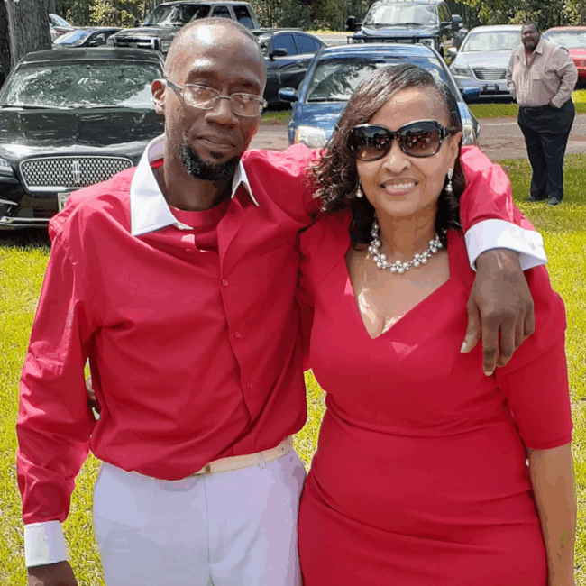 Dre Reed dressed in white slacks with a red button down shirt that has a white collar and white cuffs with his arm around his mom's shoulder. Mom is dressed in a red dress with a fancy pearl necklace and with sunglasses on.
