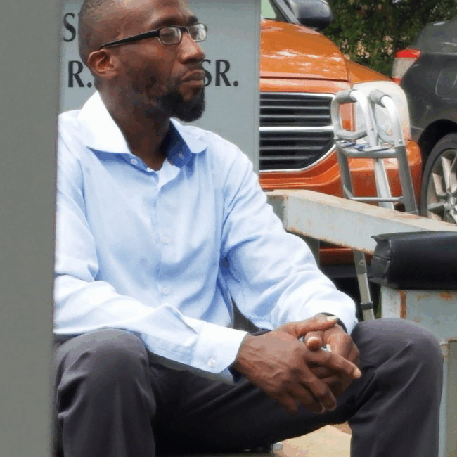 Dre sitting down outside church in a light blue button down shirt and gray slacks. He is waiting for a fellow church member with their walker nearby