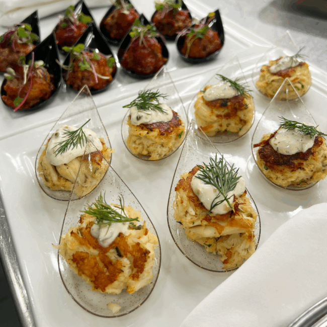 mini jumbo lump crab cakes and mini prime meatballs in small plastic tear drop shaped vessels sitting on a square white china plate