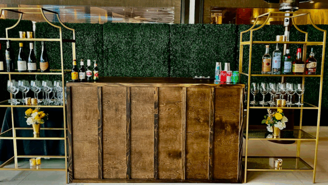 a wooden bar flanked by gold iron shelving filled with wine glasses, liquor bottles, wine bottles and fresh florals with a large greenery style hedge in the background