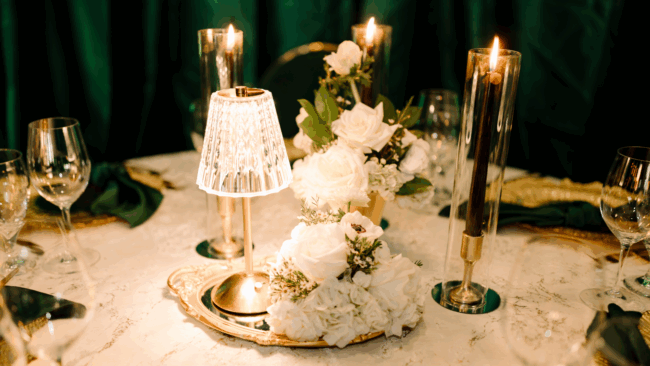 a private dining table dressed in white linen with gold and crystal table lamps, cylinder glass tapered candles, wine glasses, gold chargers and dark green napkins