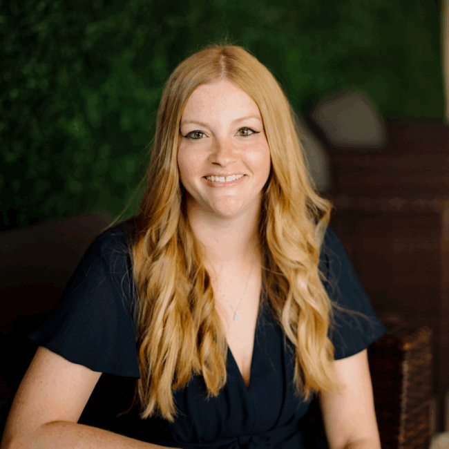 headshot of Amanda Chamber with long strawberry blond hair and wearing a navy dress sitting in a chair