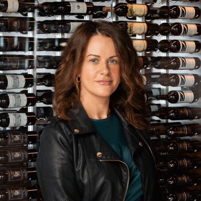 headshot of Cassandra Pancheva in a green blouse with a black leather jacket with gold adornments standing in front of the glass wine wall