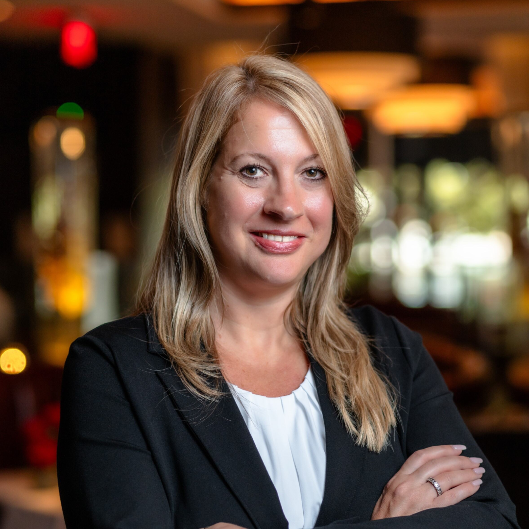 headshot of Elizabeth (Liz) Nourse in a navy suit jacket with a white blouse standing with her arms folded across her chest with the restaurant blurred in the background