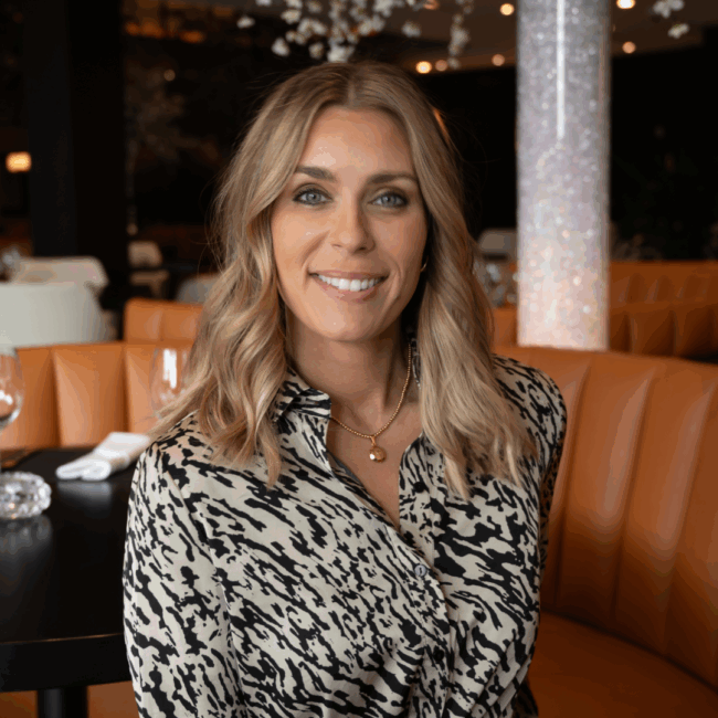 headshot of Vanessa Sbraga of Plano and Southlake in a black and cream printed dress sitting in front of the saddle tan booths with a floral tube in the background