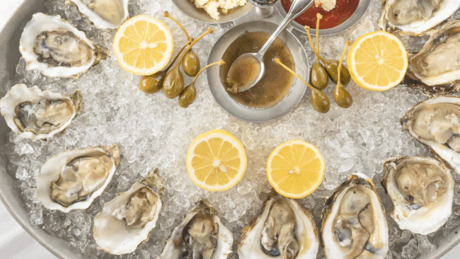 an overhead shot of freshly shucked oysters on the half shell on a large bed of ice with several lemon halves sitting in ice, migonette sauce with a spoon sitting in the dish, horseradish, cocktail sauce, and fresh caperberries