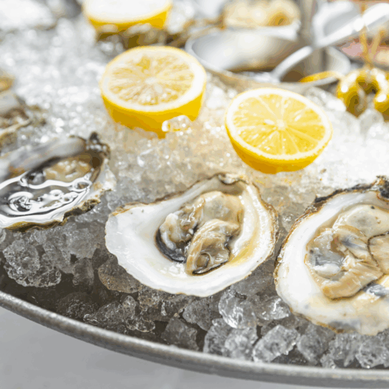 Freshly shucked oysters on the half shell on a bed of ice in a pewter tray with 2 lemon halves sitting in the ice and caper berries in the background