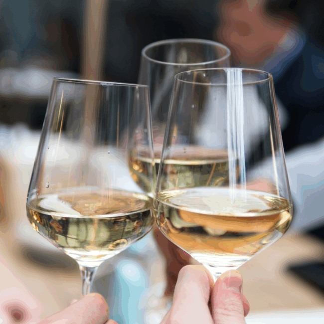 3 wine glasses filled with white wine being held by 3 hands and coming together in a toast fashion