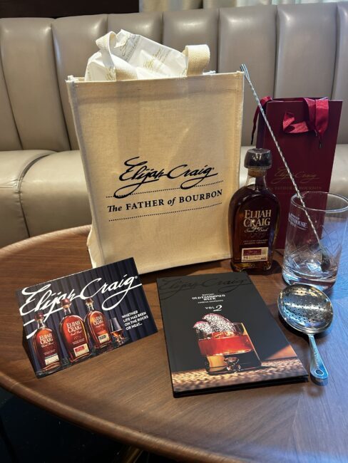 Elijah Craig Father’s Day gift set displayed on a wooden table, featuring a canvas branded gift bag, bottle of Elijah Craig Small Batch bourbon, rocks glass, cocktail mixing spoon, julep strainer, Old Fashioned recipe book, and branded gift packaging.