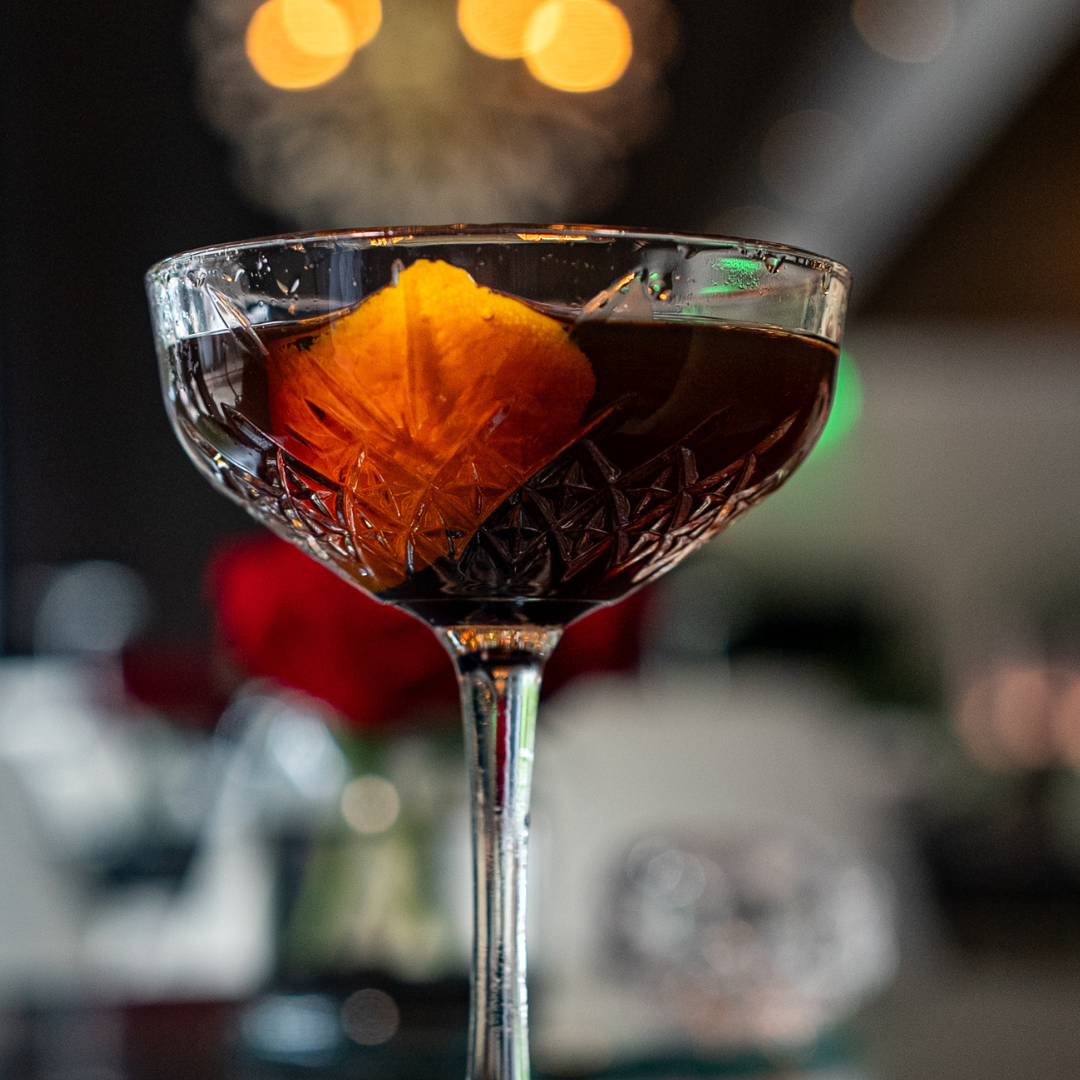 Manhattan cocktail in a crystal coupe style glass with a blurred lighted background and an orange garnish in the glass
