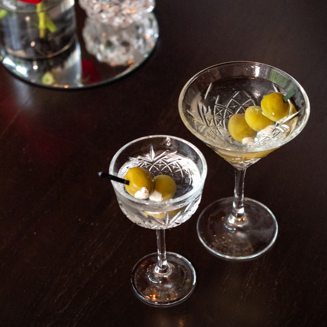 a tiny tini and regular sized dinner martini garnished with blue cheese stuffed olives in crystal stemmed glassware sitting on a dark wood table