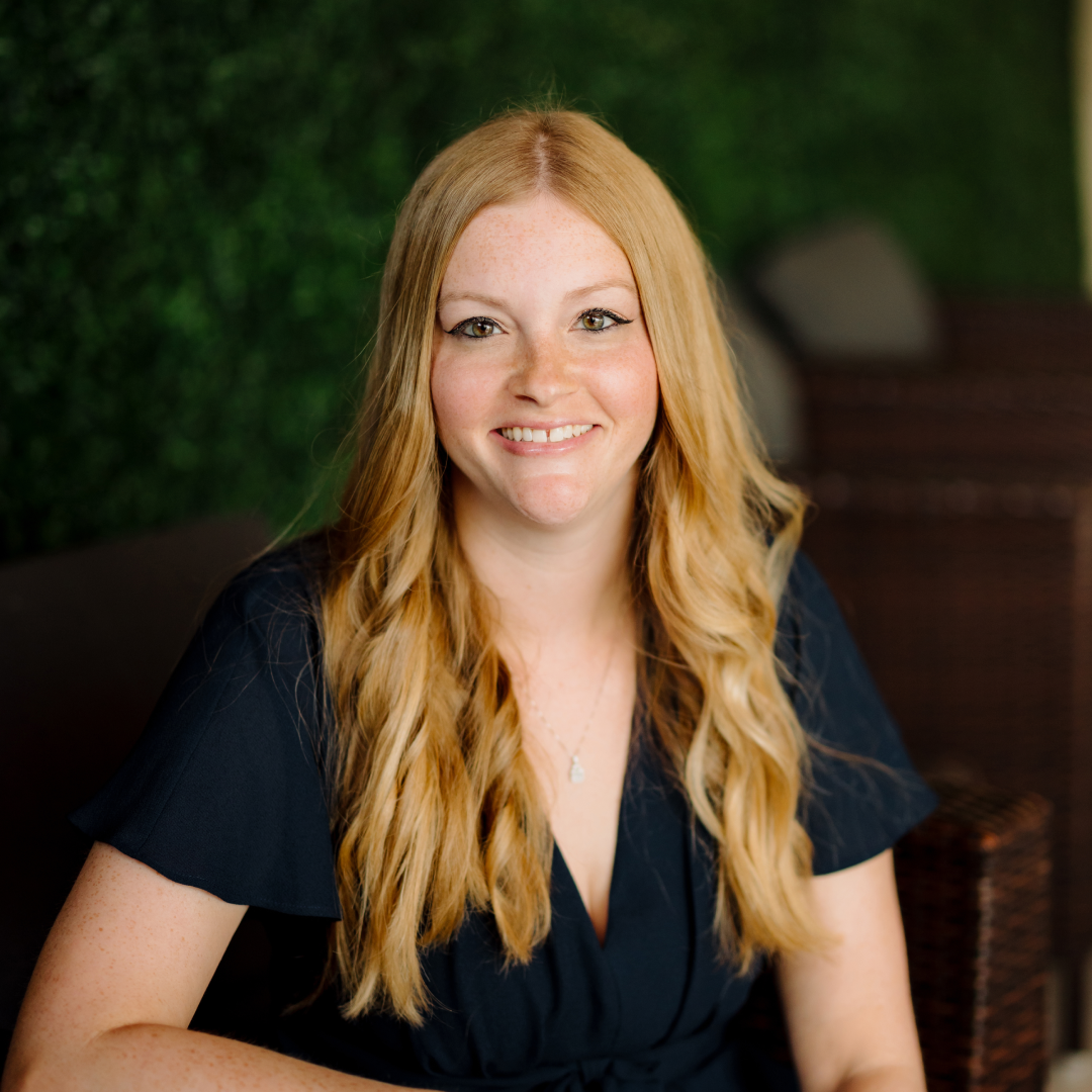 headshot of Amanda Chamber with long strawberry blond hair and wearing a navy dress sitting in a chair