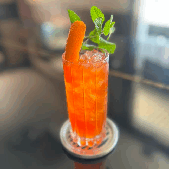 a bright orange beverage over ice in a tall collins glass with an orange peep and fresh mint garnish