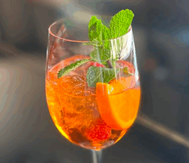 Elderflower spritz cocktail over ice in a wine glass with fresh mint, orange half wheel and raspberry