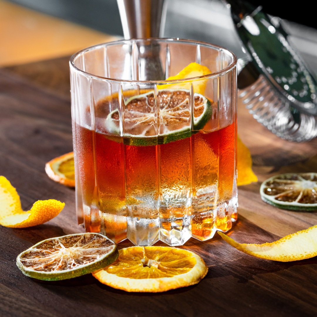 the rum fashioned is a deep orange color and served in a ridged rocks glass over a block of ice with a dehydrated lime wheel. This glass has lemon and lime wheels and twists atop a wooden table top