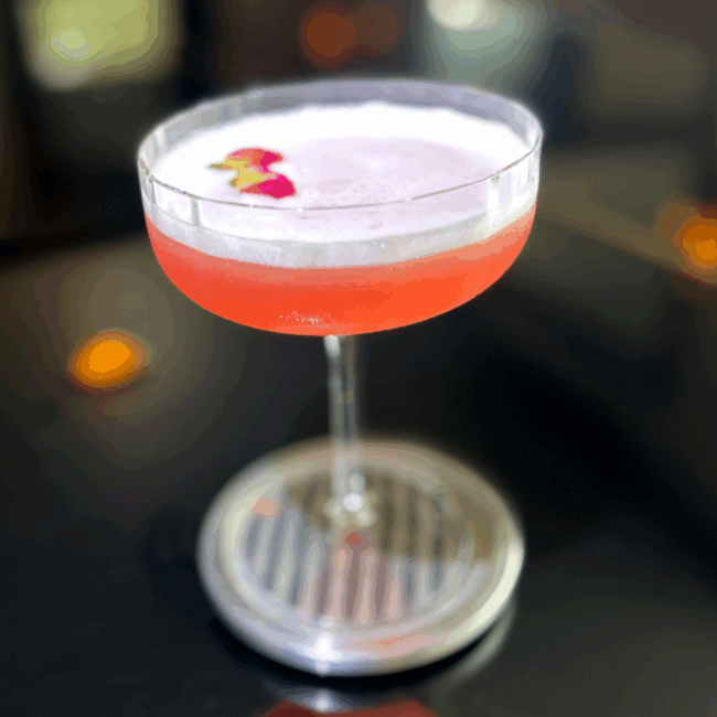 the Lucky Clover cocktail is a desert orange color with pink undertones and served in a ridged coupe glass with a slightly foamy top layer to the cocktail and garnished with two delicate mini roses.