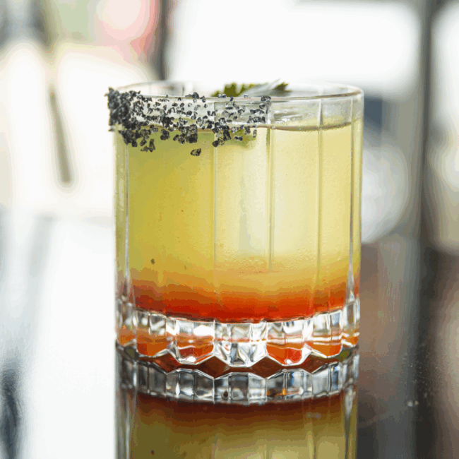 The Smoke and mirrors cocktail has a light yellow hue with orangish liqueur floating in the bottom of the glass. Covered with a lava salted rim in a rocks style glass with a green garnish
