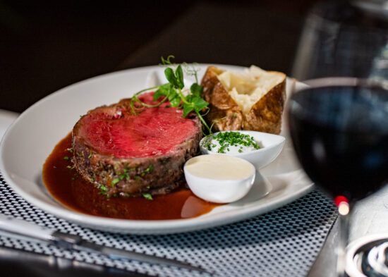 A slice of slow roasted prime rib with au jus, a baked potato and ramekin of butter on a white china plate with a glass of red wine in the foreground