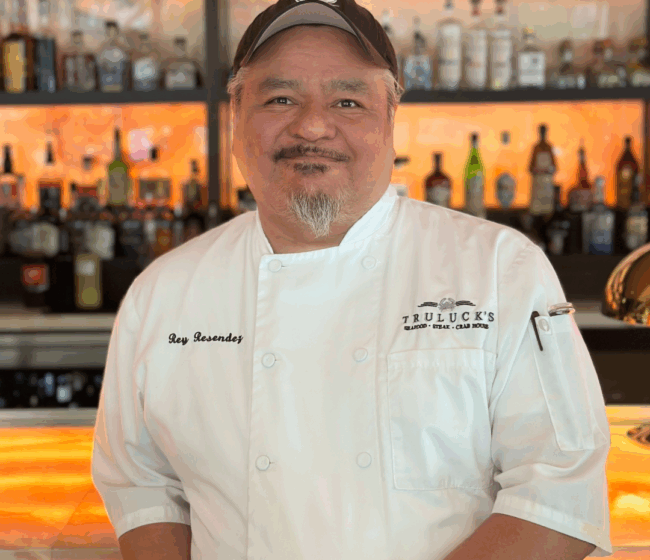 Photo of Rey Resendez in his white Truluck's chef's coat with a black Truluck's crab logo hat standing in front of the bartop with his elbow on the bar.
