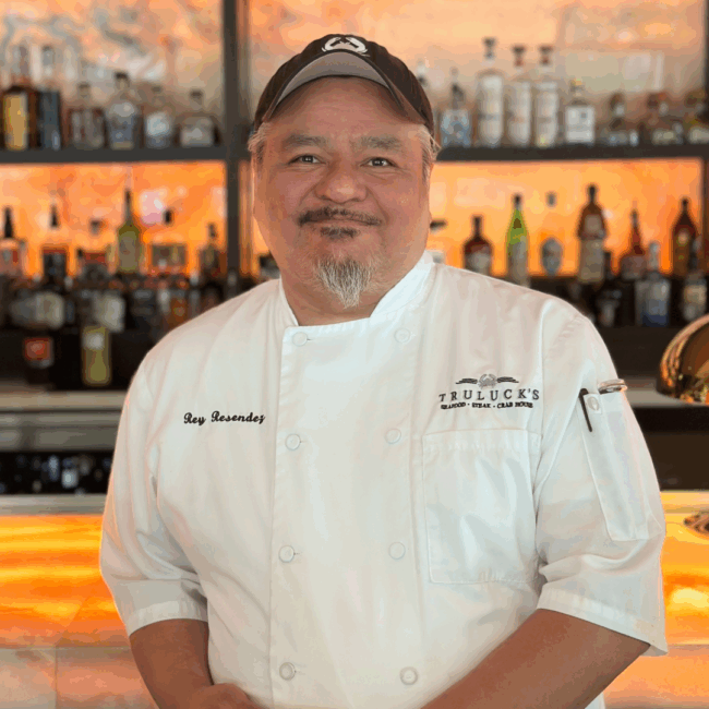 Photo of Rey Resendez in his white Truluck's chef's coat with a black Truluck's crab logo hat standing in front of the bartop with his elbow on the bar.