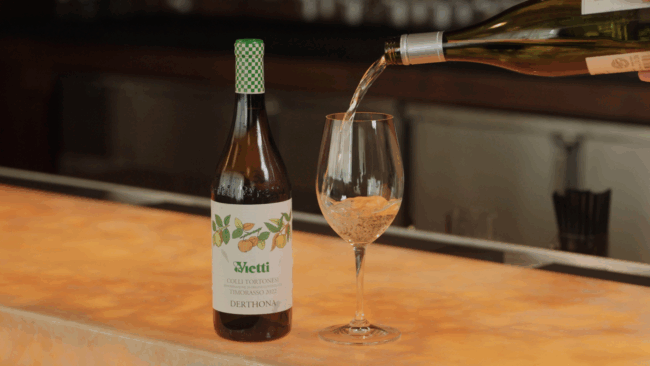 a bottle of Vietti Colli Tortonesi white wine sitting on the bartop with a white wine glass next to it being filled by another bottle of wine
