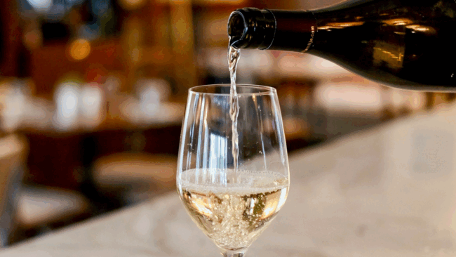 a glass of white wine being poured from the bottle while sitting on the bartop