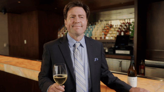 shot of beverage director, Dave Mattern standing in front of the bar in a dark gray suit with light blue shirt, striped tie and blue pocket square. He's holding a glass of white wine while smiling at someone off to the side of the camera. There is a bottle of white wine on the bar top and the back bar is filled with display bottles
