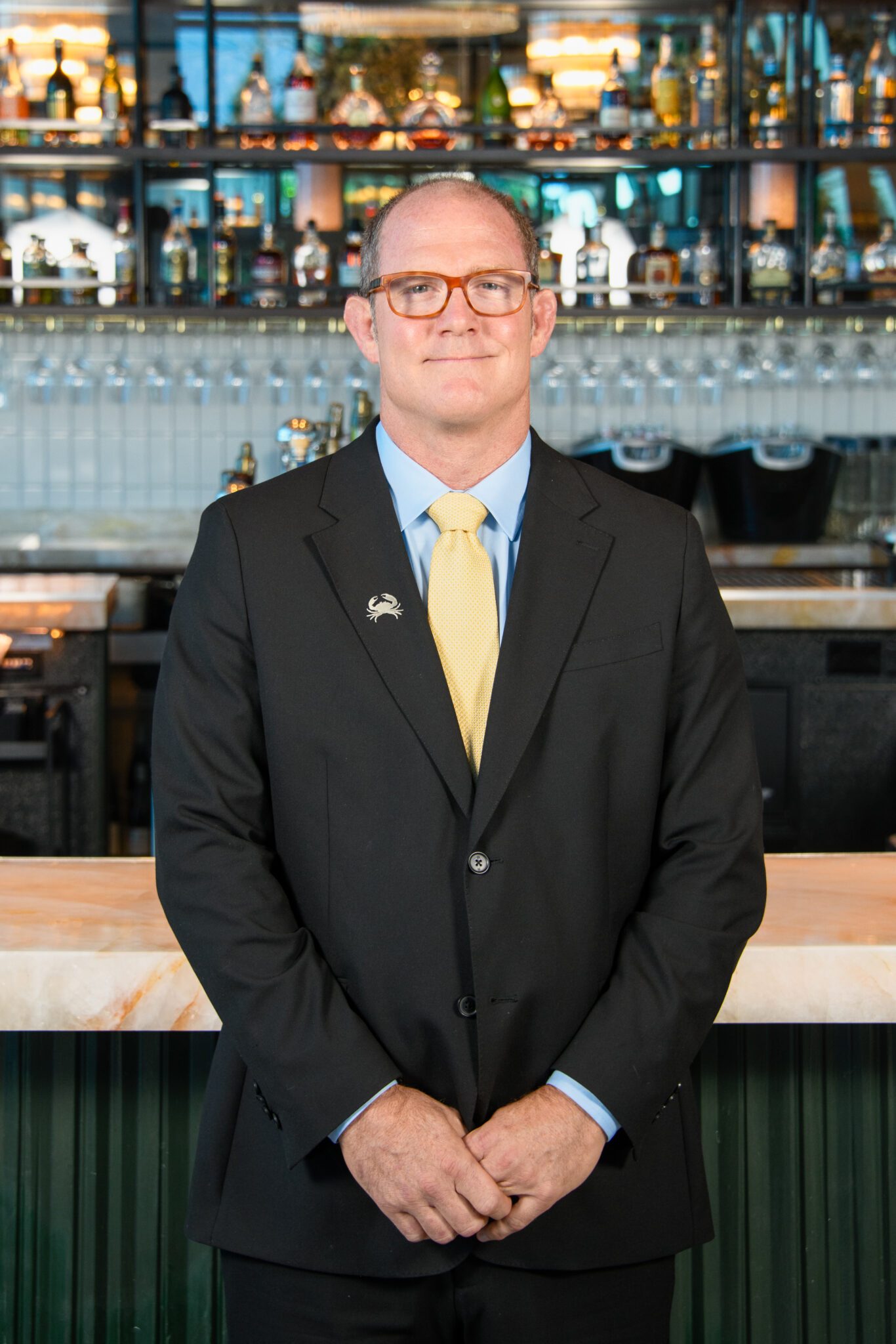 headshot of Fort Lauderdale general manager, Bill Gibson, standing in front of the bar in a black suit with a light blue shirt and yellow tie with his hands crossed in front of him.
