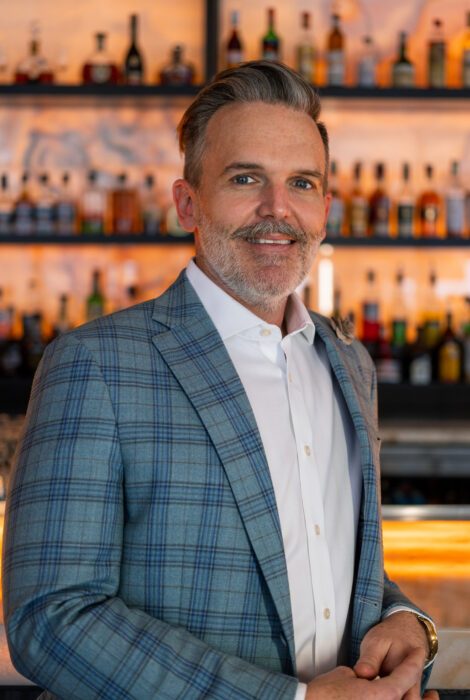 headshot of Riley Hutton operating partner in a blue plaid suit jacket with white button up shirt with his elbow leaned on the bar top. He is smiling and looking at the camera with a beared face.