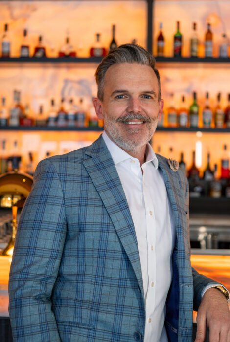 Riley Hutton, operating partner headshot. He is in a blue plaid sport coat with white button up shirt and has his elbow leaned on the glowing bartop. The backlit back bar filled with liquor bottles is in the background.