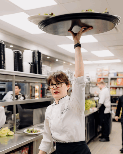 A female Truluck's server in a white chef coat in the kitchen holding a tray of food way up over her head with her arm extended high