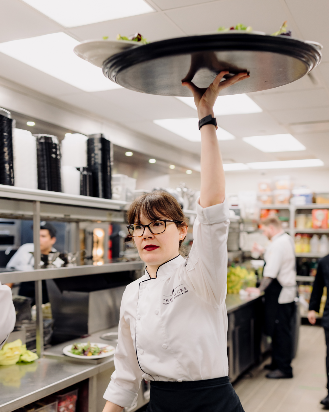 A female Truluck's server in a white chef coat in the kitchen holding a tray of food way up over her head with her arm extended high