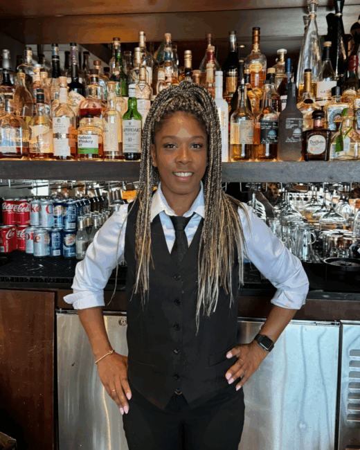 Darsha our bartender in Truluck's Rosemont in a bar uniform that includes a black vest with shirt and tie. She has blond braids pulled in a ponytail and is standing in front of the back bar area.