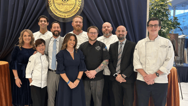 Team Woodlands standing together in front of a navy draped backdrop at the Hometown Heroes event. The team is dressed in their best and a few are in white server coats.