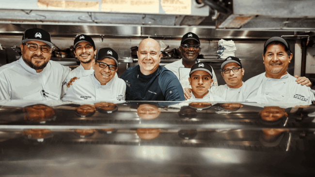 Corporate Chef Thomas and Culinary Director Chef Sammy with the Houston line cooks and Executive Chef standing on the kitchen line and smiling while looking through the stainless steel expo window