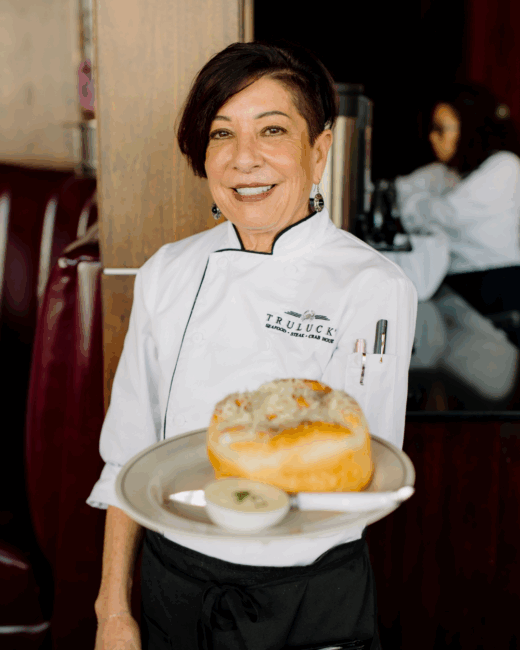 server Celeste from The Woodlands in a white long sleeved server coat with the Truluck's logo on one side. She's holding a plate of Parker House rolls with butter and smiling while standing in the dining room.