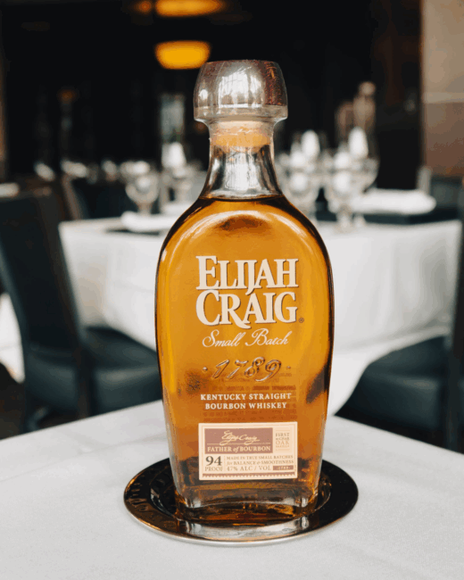 A bottle of Elijah Craig small batch 1789 Kentucky straight boubon whiskey sitting on a silver coaster on a table covered in a white table cloth