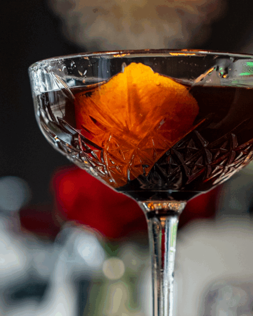 A Manhattan cocktail in a crystal stemmed coupe style glass with a large orange peel in the glass of whiskey